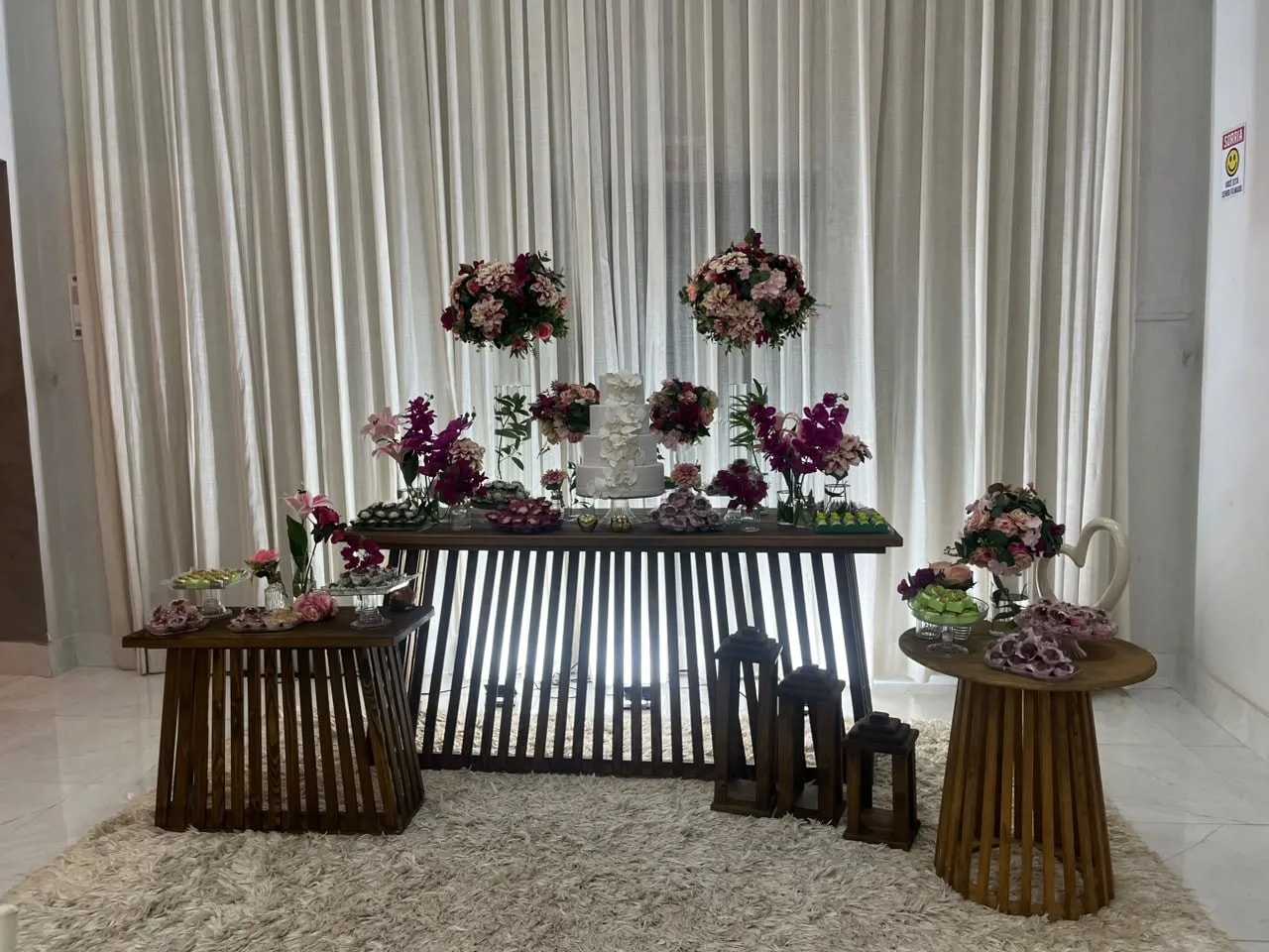 Mesa de bolo com arranjos em rosa e vinho diante de cortinas brancas